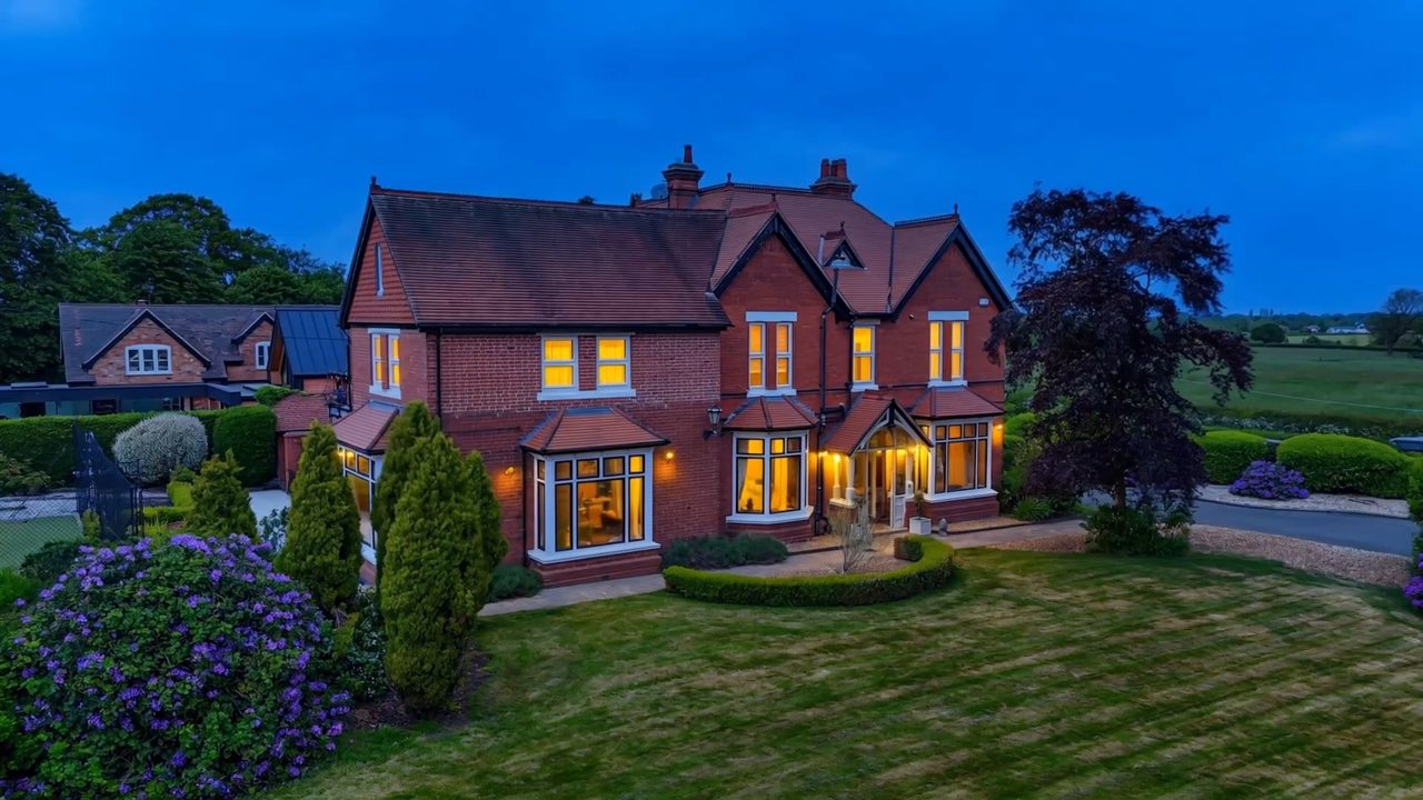A red-brick country house shown at dusk with every window lit warm.